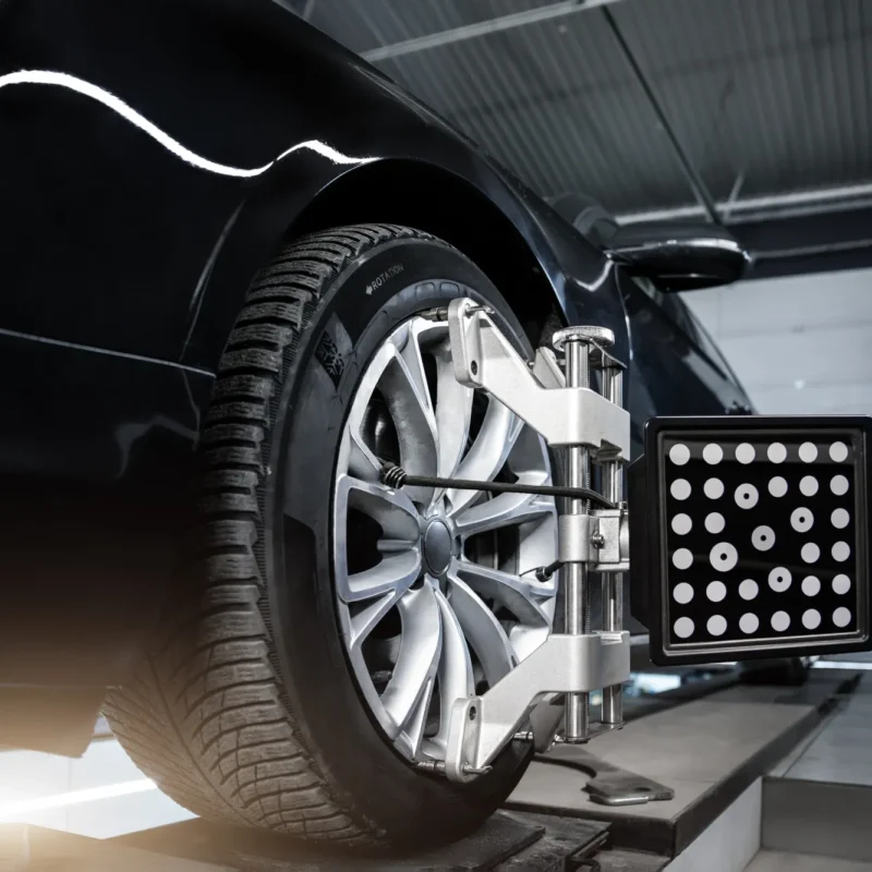 A wheel alignment target board clamped to the alloy wheel of a black car on an alignment rack in a repair shop.